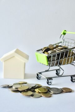 A model house standing alongside a scattered stack of coins and a small shopping cart filled with money, demonstrating concepts of property investment, real estate market costs, and housing budgets.