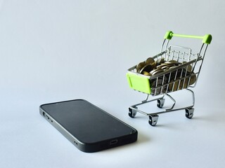 Modern smartphone lying beside a mini shopping cart packed with coins on a clean white surface, symbolizing mobile shopping apps, digital payment methods, and online financial management. © Akram