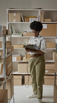 Vertical video of young adult African American woman scanning barcodes on boxes using tablet and barcode reader in warehouse. Footage ideal for e-commerce and logistics marketing