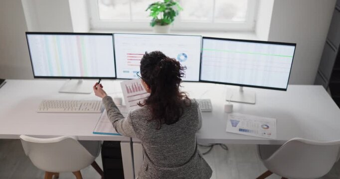 Woman analyst reviews spreadsheet data report on computer. Three monitors show accounting and finance figures in a clean office, with charts and KPIs. Clear data driven financial audit concept.