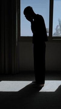 Silhouette of a distressed man standing alone by a window and covering his face, overwhelmed by emotional stress, worry and a difficult realization in a dark dramatic room, vertical