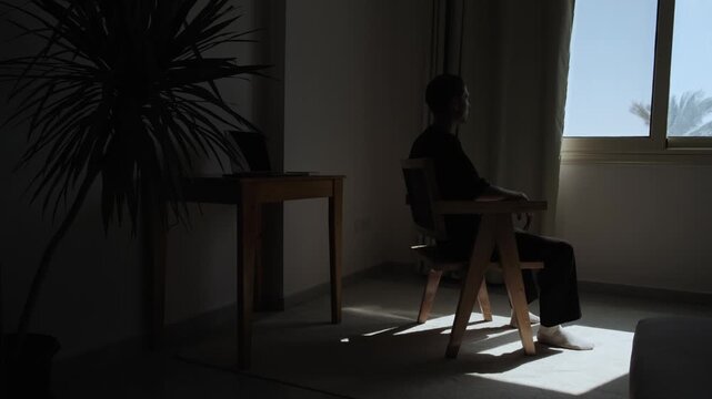 Silhouette of lonely man sitting alone by a window in a dark room, experiencing isolation, sadness and emotional loneliness