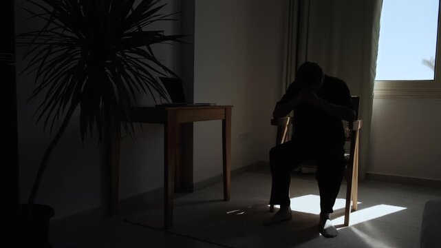 Silhouette of lonely man sitting alone in dark room, lowering his head into his hands in emotional distress, expressing sadness, despair, isolation and inner struggle
