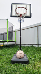 Outdoor basketball hoop with ball on grassy court beside trampoline. Recreational sport equipment for shooting practice, backyard fitness, training and active summer lifestyle. © Анна Маркина