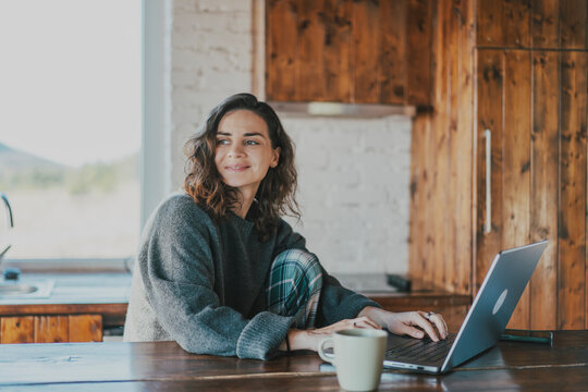 Millennial woman in cozy sweater working on laptop while sitting at wooden table with coffee cup at home.