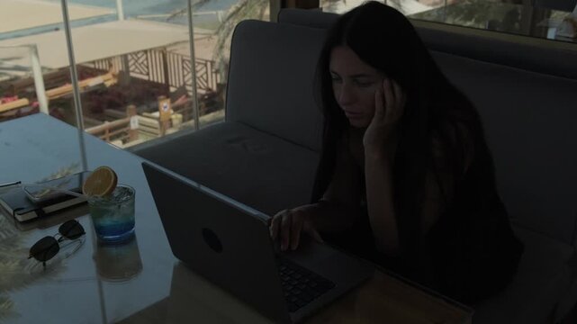 Female digital nomad working on laptop in tropical resort cafe while people relax outside, palm trees and vacation atmosphere showing work and travel lifestyle