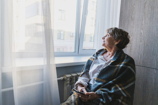 Lonely senior Caucasian woman sitting by a radiator near the window, wrapped in a blanket with a smartphone, conceptualizing pensioner isolation.
