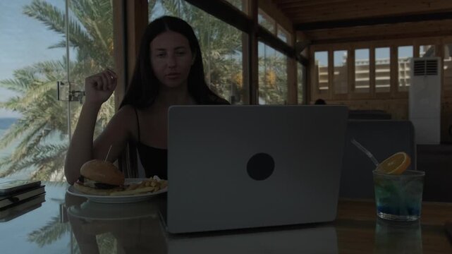 Woman eating fast food while working on laptop at seaside cafe showing unhealthy work habits sedentary lifestyle poor work life balance convenience food and lack of breaks