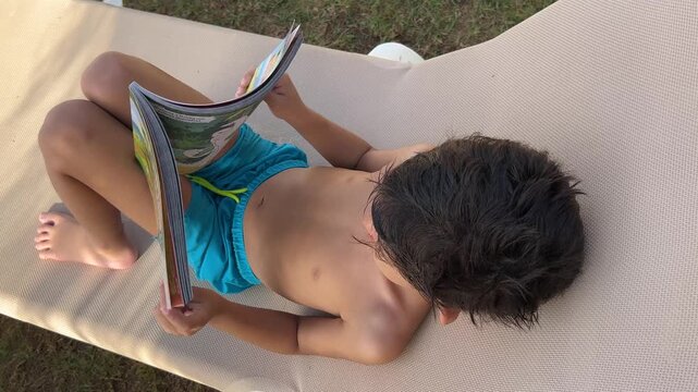 Static top-down shot of a young boy in blue swimming trunks lying relaxed on a pool deck chair while reading a book during his summer vacation.