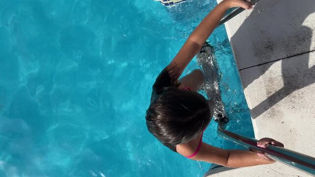 Static top-down shot of a young girl in a pink swimsuit climbing the metal ladder to exit the crystal clear water of the swimming pool on a sunny summer day.