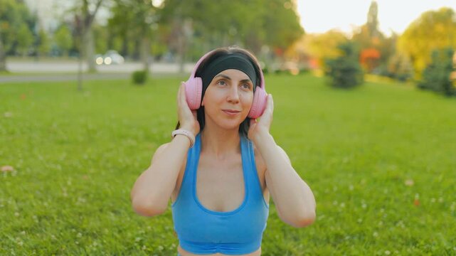 Young athletic woman listening to music with pink headphones in the sunny park.