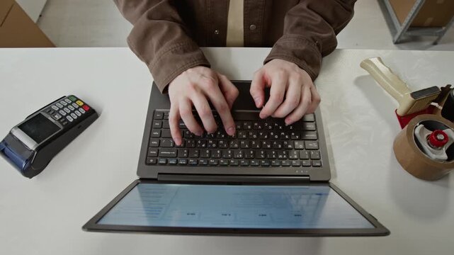 Young adult Caucasian man typing on laptop at desk with payment terminal and barcode scanner. Clip is perfect for e-commerce, retail checkout, and small business logistics videos