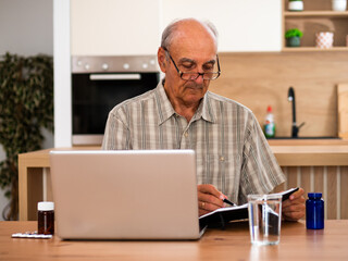 Fototapeta premium Senior man using laptop to read about prescription medicine while writing notes and checking information about his health at home.