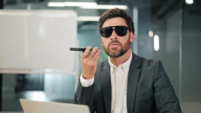 Professional in dark sunglasses speaks into smartphone for voice message or assistant at office desk. Modern business communication uses mobile technology for efficient work.
