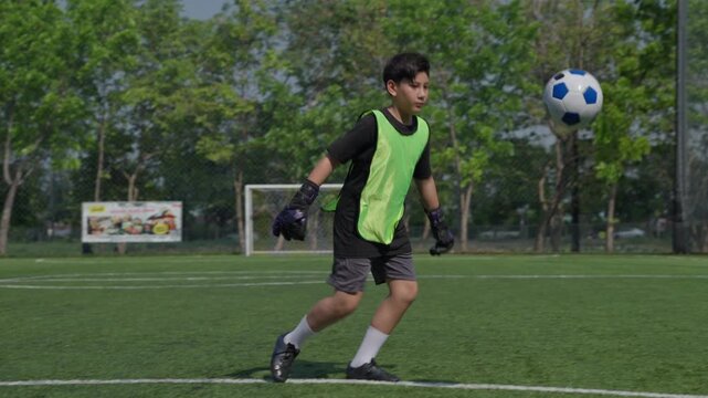 Young male soccer player green training vest controls and kicks ball during outdoor practice artificial turf, focused footwork session surrounded by trees, fencing, sunny park, goal equipment