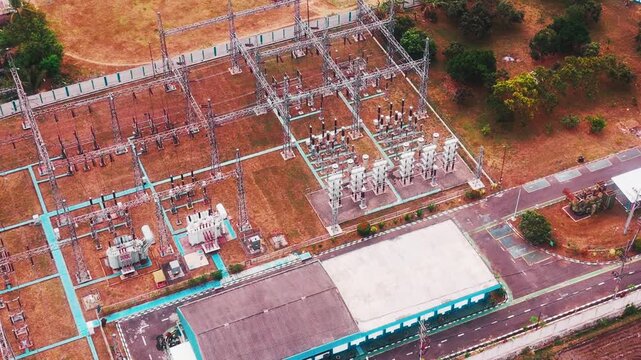 Aerial drone view of a large electrical substation featuring high voltage transformers, transmission towers, steel structures, switchgear, and power distribution equipment surrounded by dense urban bu