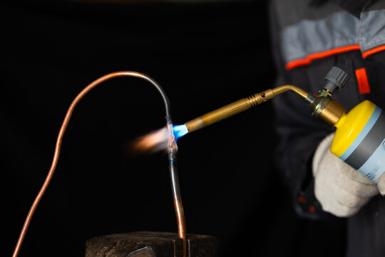  a worker in protective gloves brazing a copper pipe with a gas torch. Professional plumber soldering thin copper tubing with a hot blue flame in a workshop, plumbing and HVAC installation concept.