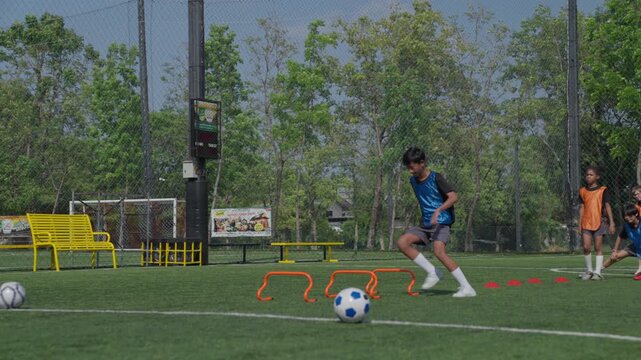 Youth Soccer Player Dribbling Ball Through Agility Hurdles During Outdoor Training on Artificial Turf Field with Coach and Teammates Practicing Skills and Ball Control