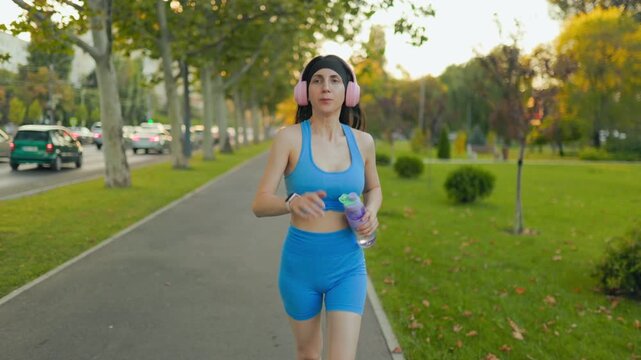 Active woman running on the park path with headphones and water bottle.