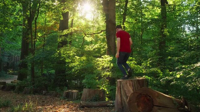 Young man playfully jumping on logs in a sunny lush green forest, with the beautiful sun in the background