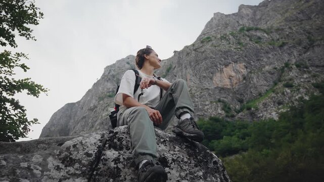Young female traveler with a backpack sitting on a large rock and enjoying a breathtaking mountain view, capturing freedom, adventure, relaxation and connection with nature.