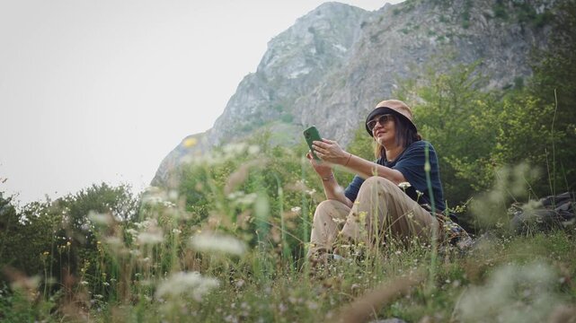 Female traveler using a smartphone while sitting in a scenic mountain meadow with majestic mountains in the background, capturing connectivity, navigation and modern adventure travel in nature.