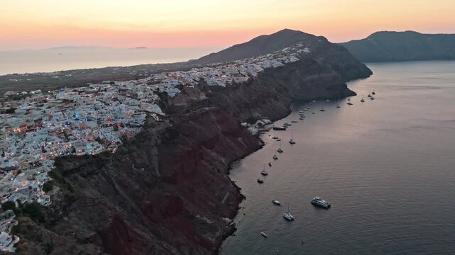 Long row of anchored boats sits below the rising cliffs of Oia Santorini as the town lights glow against a warm dawn sky.