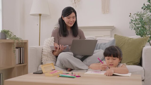 Asian mother helping girl child with homework on laptop beside sofa at home, warm family learning with woman and child drawing, cozy living room connection during relaxed creative afternoon activity