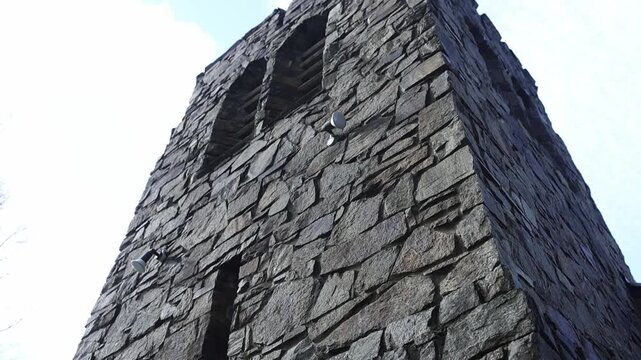 Dramatic upward wide shot of an old stone church tower in Olinda, Melbourne, set against a bright, cloudy sky. Textured masonry and weathered details highlighted by natural daylight.