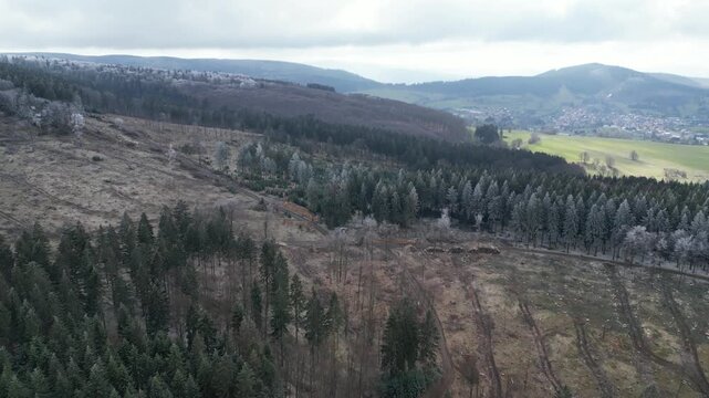 Drone footage over a deforested slope and coniferous forest, leading to a small village nestled in the Thuringian hills of Germany. Cloudy winter day with a peaceful, remote landscape.