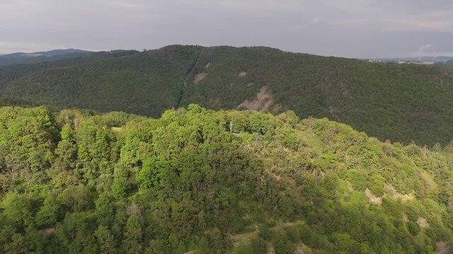 An aerial drone shot of dense forested hills in Rhineland-Palatinate, Germany. The camera pulls back and booms up, revealing a prominent cross on a serene hilltop. Peaceful and remote nature scenery.
