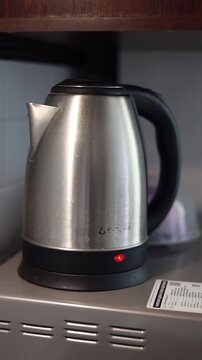 Stainless steel electric kettle with black handle and red power indicator light, standing on a kitchen shelf beneath wooden countertop