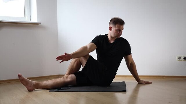 Man sitting on yoga mat performing seated forward bend to stretch his hamstrings, exercising at home in bright empty room