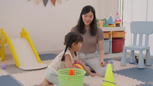Asian woman clapping beside child playing colorful toy hoop bright playroom, family creative learning activity with mother and daughter, joyful childhood fun near slide, chair, storage bins