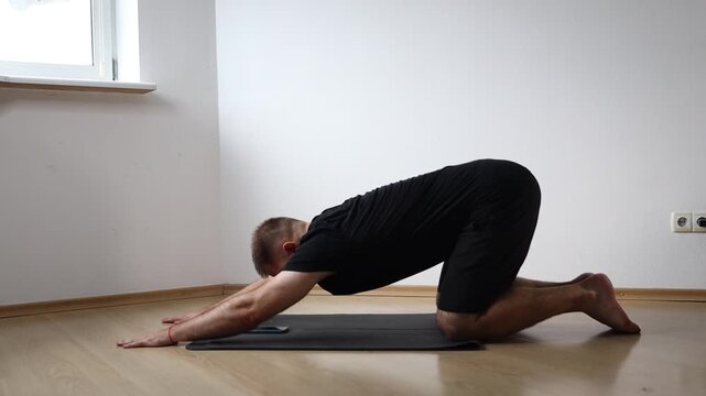 Man sitting on yoga mat performing seated forward bend to stretch his hamstrings, exercising at home in bright empty room