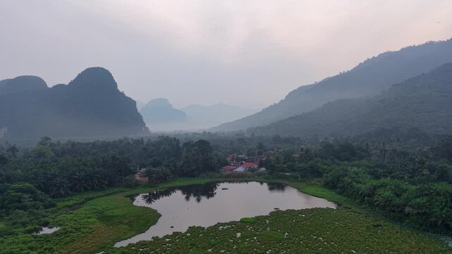 Misty karst mountains loom over a serene landscape. Calm lake reflects the dense forest surroundings at Kampar, Perak