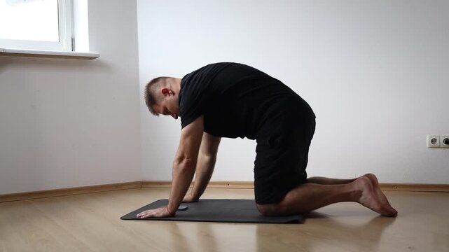 Man sitting on yoga mat performing seated forward bend to stretch his hamstrings, exercising at home in bright empty room
