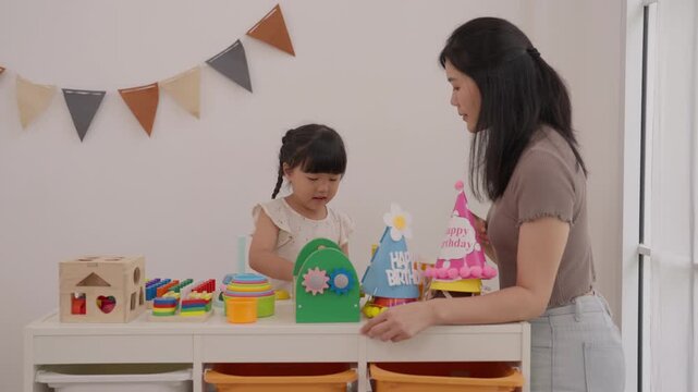 Asian mother, woman and child daughter birthday celebration with colorful toy arrangement and cake at home, joyful family moment featuring mother and daughter sharing festive happiness together