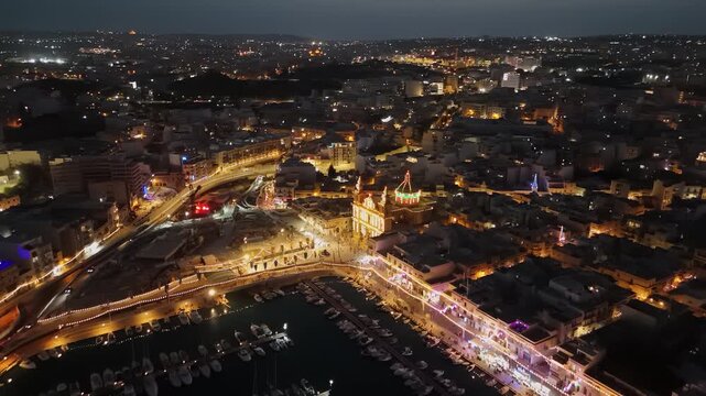 Msida, Malta marina and waterfront skyline with illuminated boats, church and apartment buildings at dusk, coastal harbor traffic and public square seen in aerial view.