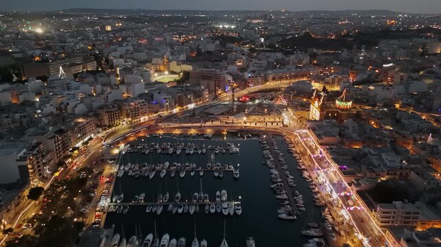 Msida, Malta, Knisja Parrokkjali tal-Imsida and marina during a village festa at dusk, with illuminated waterfront, boats, traffic, and coastal cityscape in aerial view.