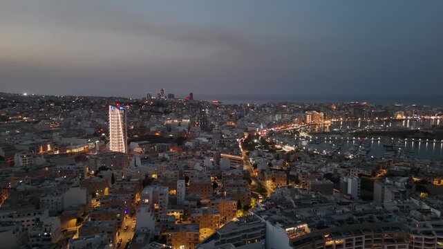 Ta' Xbiex, Malta marina with boats and yachts along the Mediterranean waterfront at dusk, featuring city lights, modern apartments and office buildings in a wide drone view.