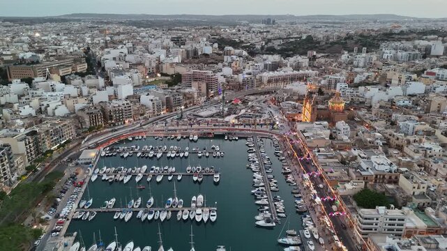 Msida, Malta waterfront and Knisja Parrokkjali tal-Imsida during a village festa with festive lights, boats and city skyline along the harbor at evening, captured by drone.