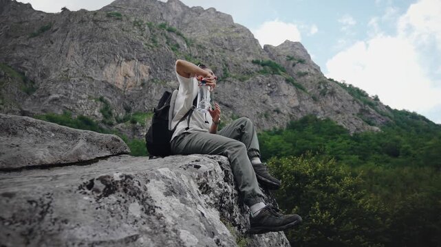 Young female traveler with a backpack drinking water from a reusable bottle while sitting on a rock and enjoying a scenic mountain view during a hiking adventure.