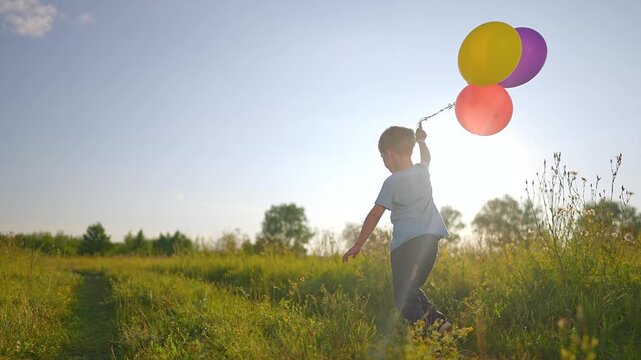 Boy runs through meadow holding colorful balloons. Child plays with balloon bundle in field. Kid running at sunset. Boy holds balloons while playing. Child runs with colorful balloon outdoor.