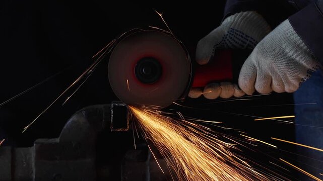  a worker in protective gloves cutting a metal square pipe with an angle grinder in 4K slow motion. Stream of bright orange sparks flying from the abrasive disc against a dark background