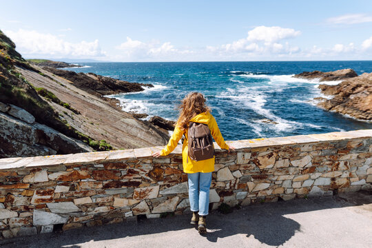 Hiker in yellow raincoat and with laptop enjoys view of ocean near a lighthouse. Woman in bright raincoat feels free by ocean at sunset. Concept of hiking, nature, and enjoyment.