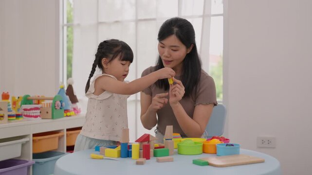 Asian mother and daughter playing colorful wooden blocks and toy food home, sharing joyful creative learning activity near bright window light, family bonding, preschool childhood education