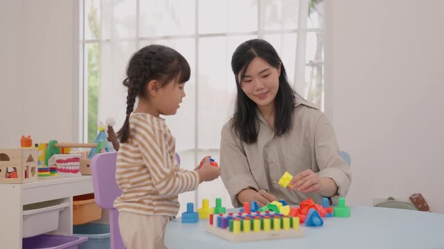 Mother and daughter building colorful block tower home, Asian family creative play with adult helping child construct toy blocks near bright window, joyful educational activity indoors for warm