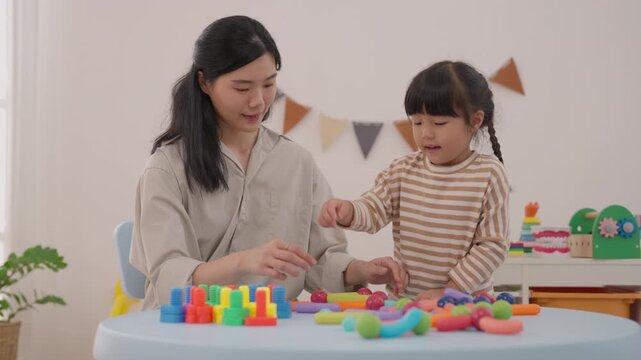 Asian mother and daughter family child woman playing colorful toy blocks home, joyful educational bonding activity with puzzle pieces, festive bunting, indoor plant, creative learning and warm