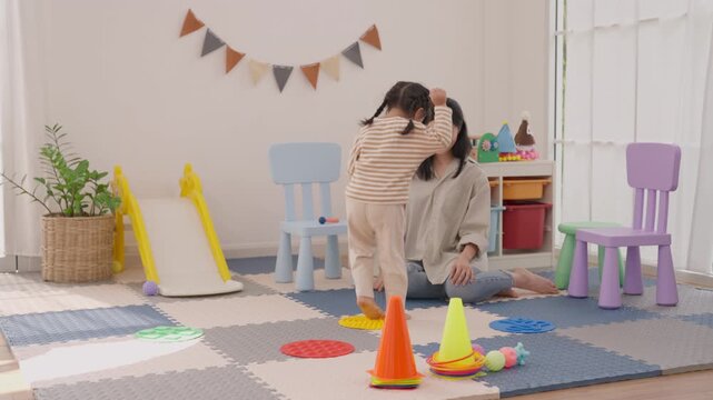 Asian mother and daughter family playroom activity with colorful toy cone, indoor slide, cheerful soft floor mat, bright furniture, festive bunting, and relaxed parent child playtime at home